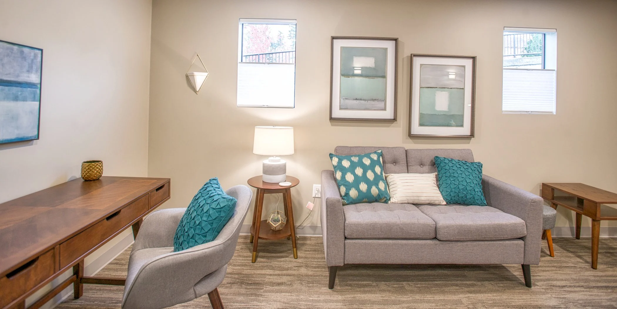 Therapy Room A modern seating area with a gray loveseat, teal throw pillows, light wood floors, framed artwork, and a wooden desk.