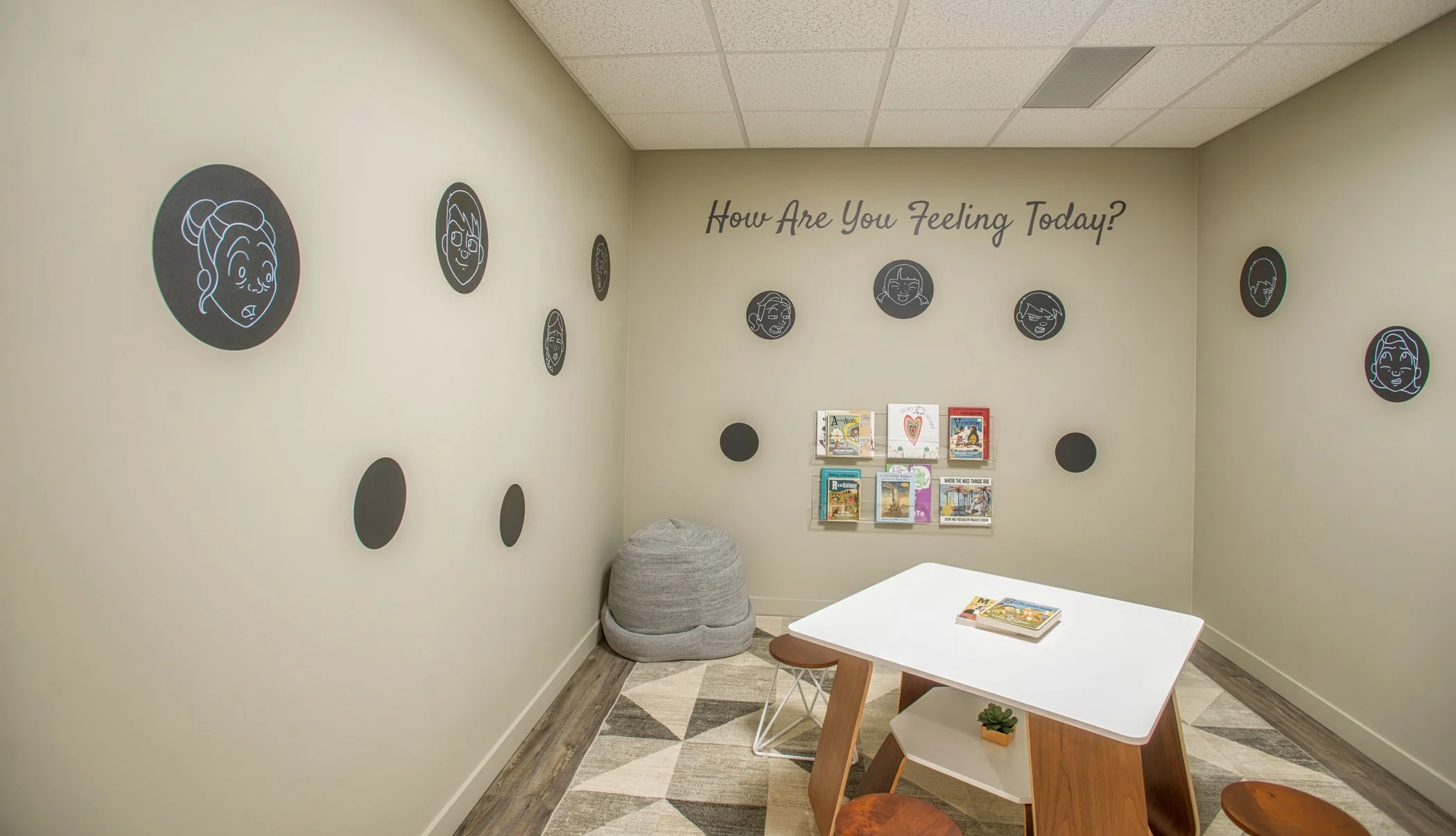 Kids waiting room A children's therapy room with a wall decal reading 'How Are You Feeling Today?' surrounded by circular emoji illustrations, a small activity table, a beanbag chair, and a book display.