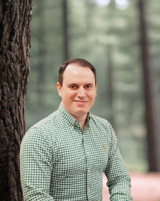 David Cueto An outdoor headshot portrait of a smiling man posing next to a textured tree trunk in a softly focused wooded area.