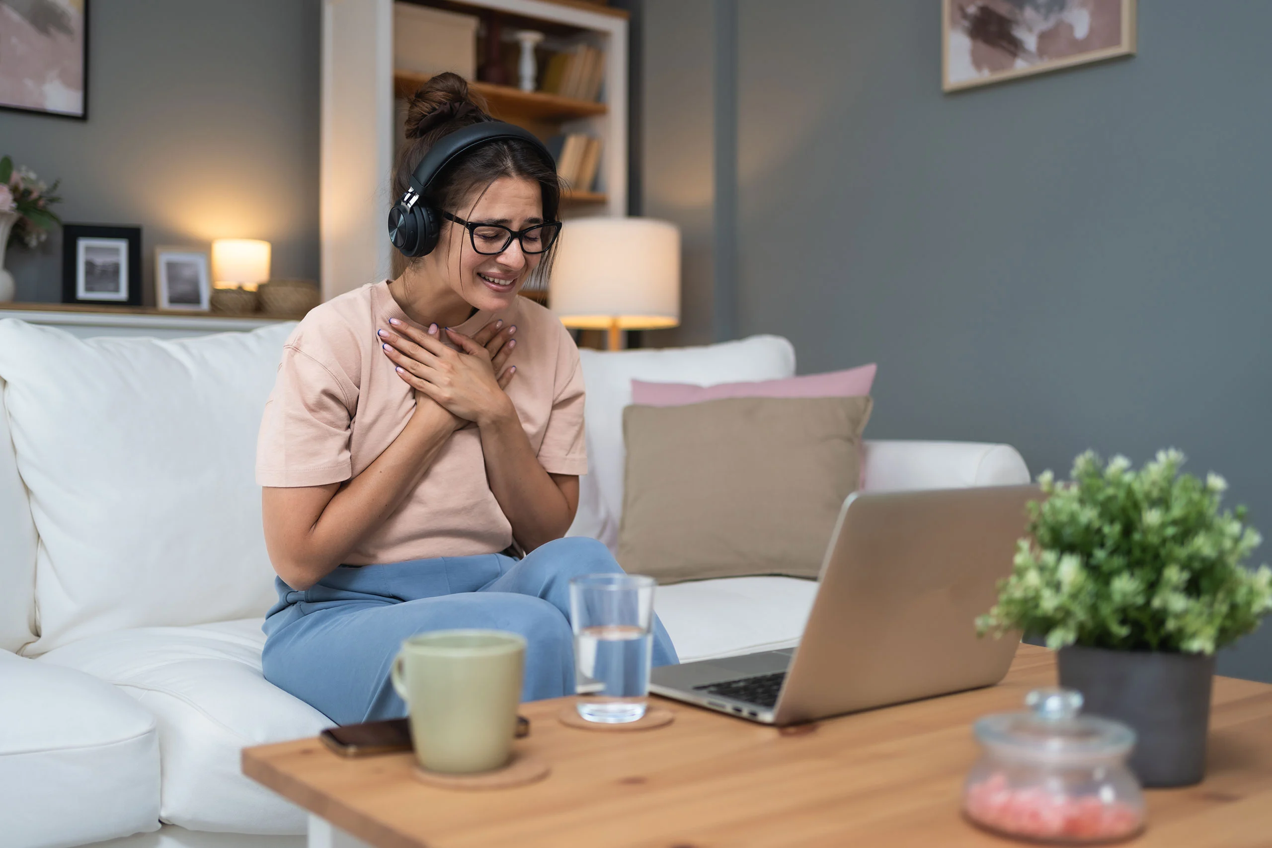 A woman wearing glasses and headphones sits on a couch, smiling emotionally with her hands over her heart during a video call on her laptop.