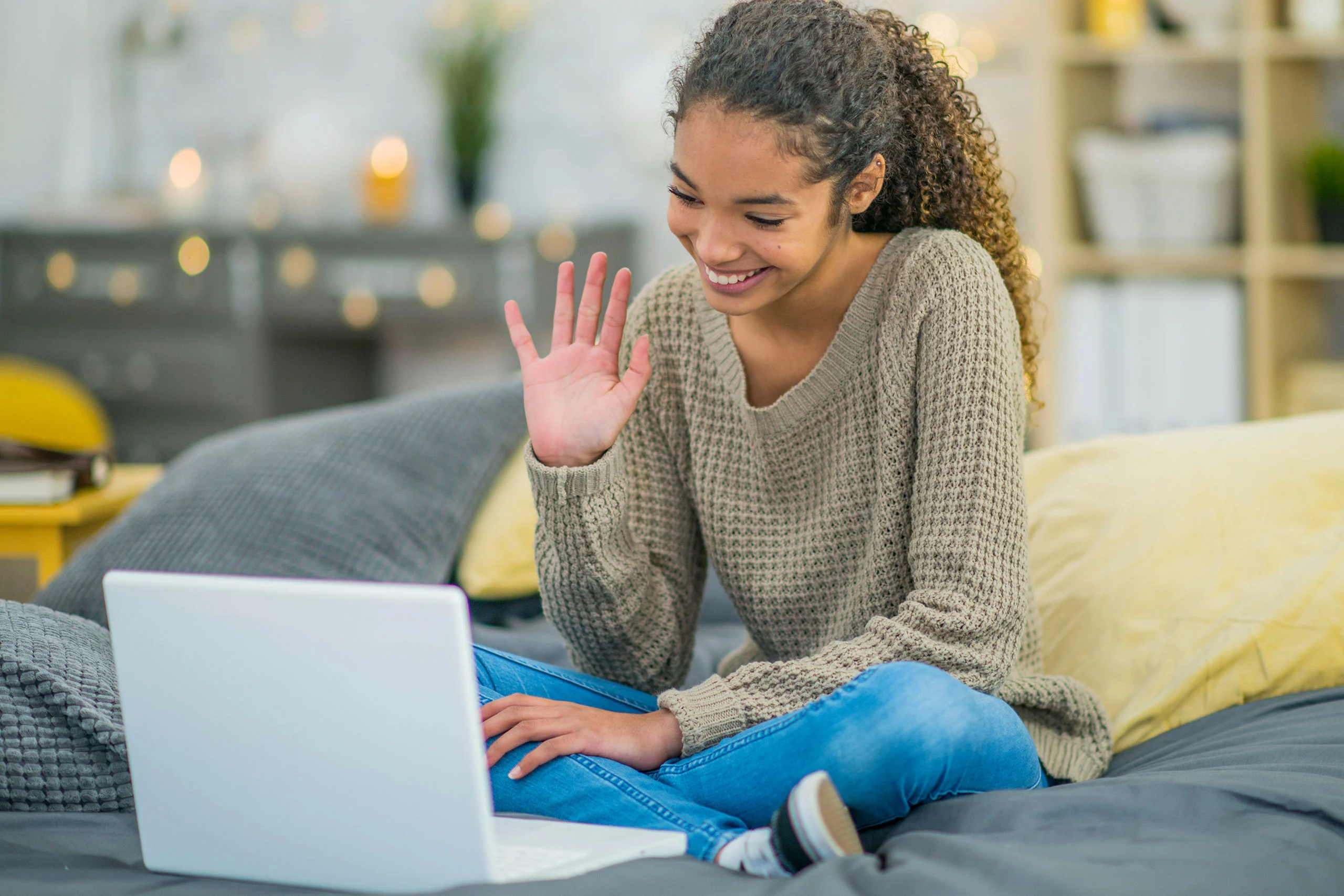 Group Therapy – One – Centered Connections A smiling teenage girl sitting cross-legged on a bed, waving hello during a video call on her laptop.
