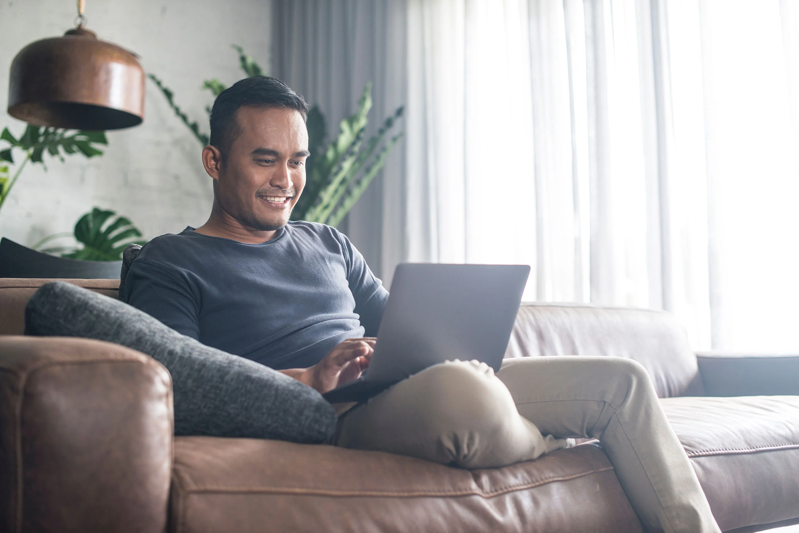 Group Therapy – Two – Centered Connections A smiling man sitting comfortably on a leather sofa while using his laptop for a video call.