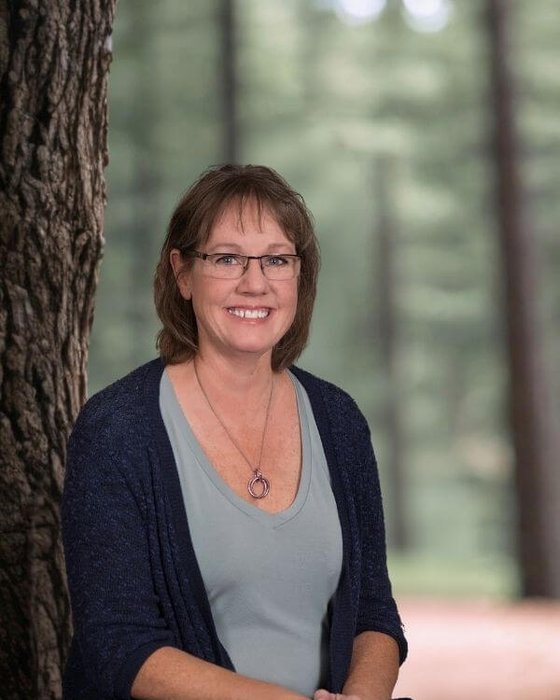 Janice An outdoor headshot portrait of a smiling woman with glasses and medium length brown hair in a softly focused wooded area.
