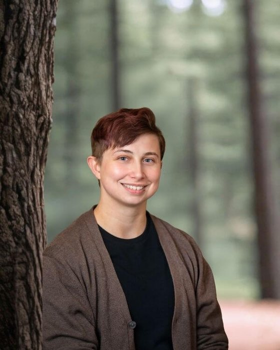 Joseph Kalita A smiling person wearing a brown cardigan outdoors in a forest.