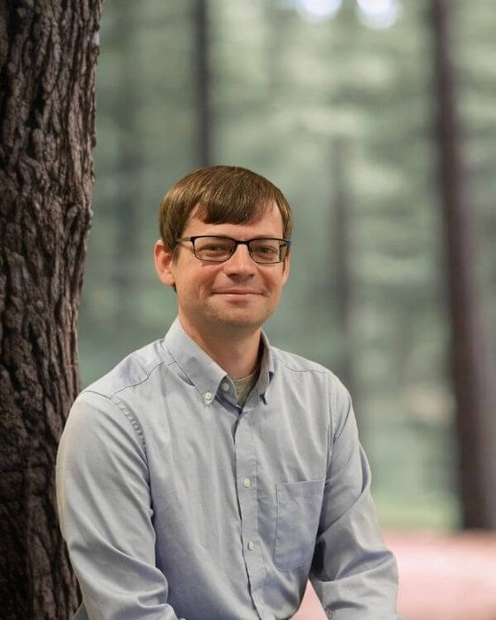 Michael Viveiros An outdoor headshot portrait of a smiling man with glasses in a softly focussed wooded area.