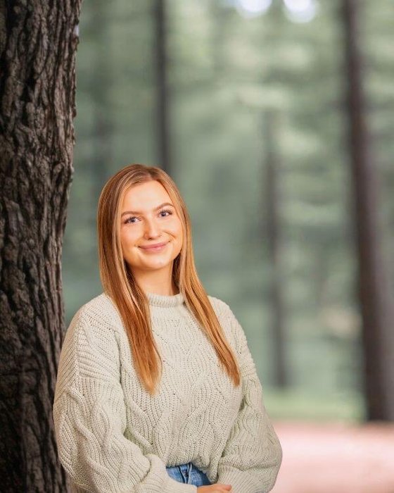 Riley Mills An outdoor portrait of a smiling woman posing next to a large tree trunk in a softly focused wooded setting.