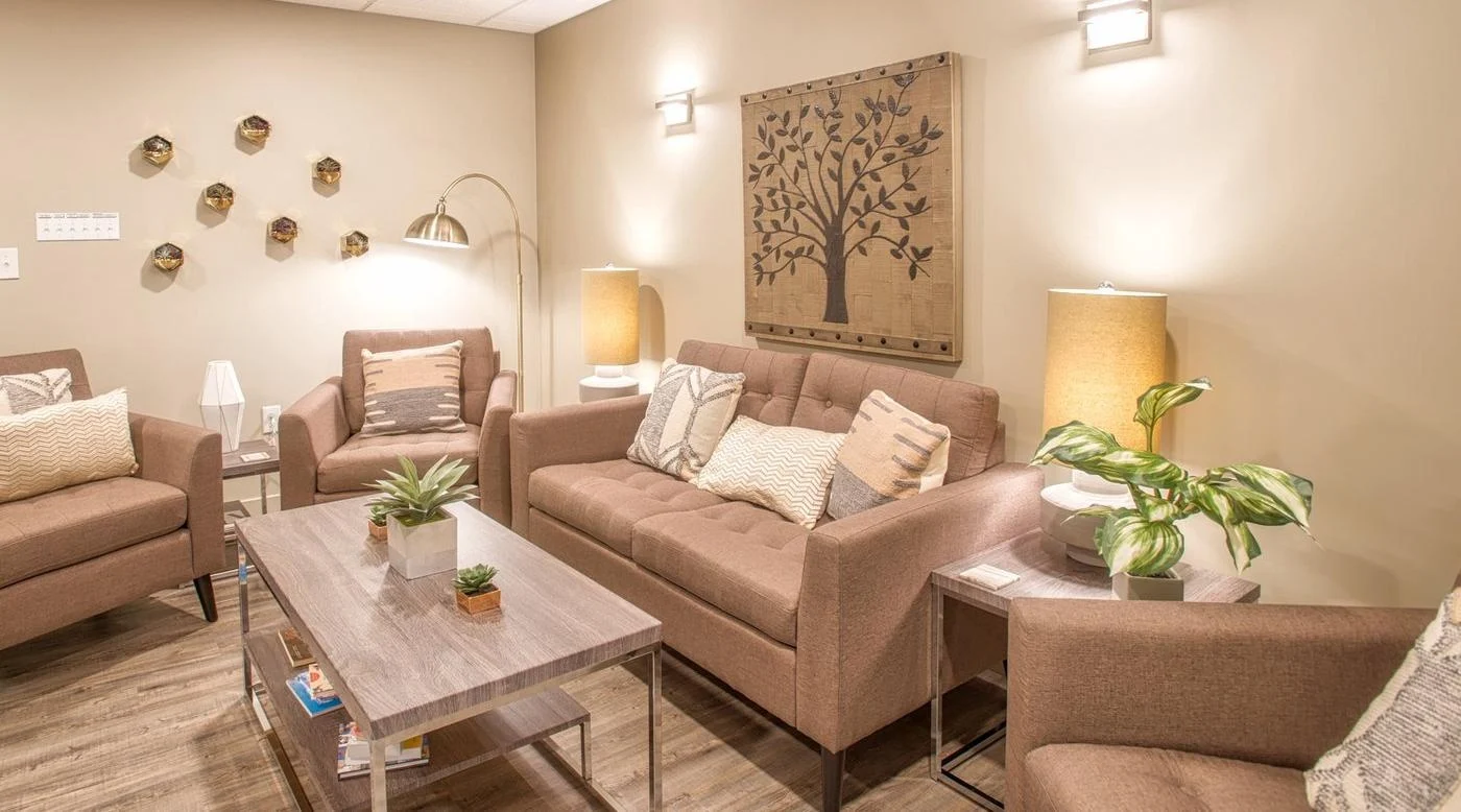 Waiting Room – Cropped – Centered Connections A modern seating area with a beige furniture, throw pillows, light wood floors, framed artwork, and a coffee table.