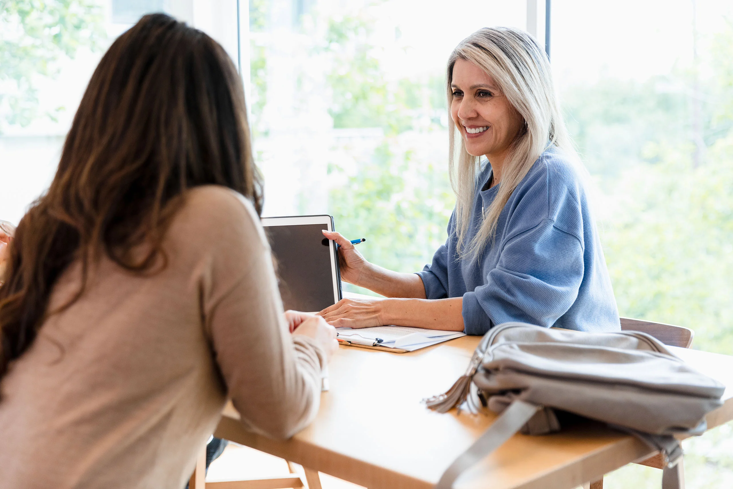 A smiling blonde woman sitting at a table and showing a digital tablet to another woman during a meeting.