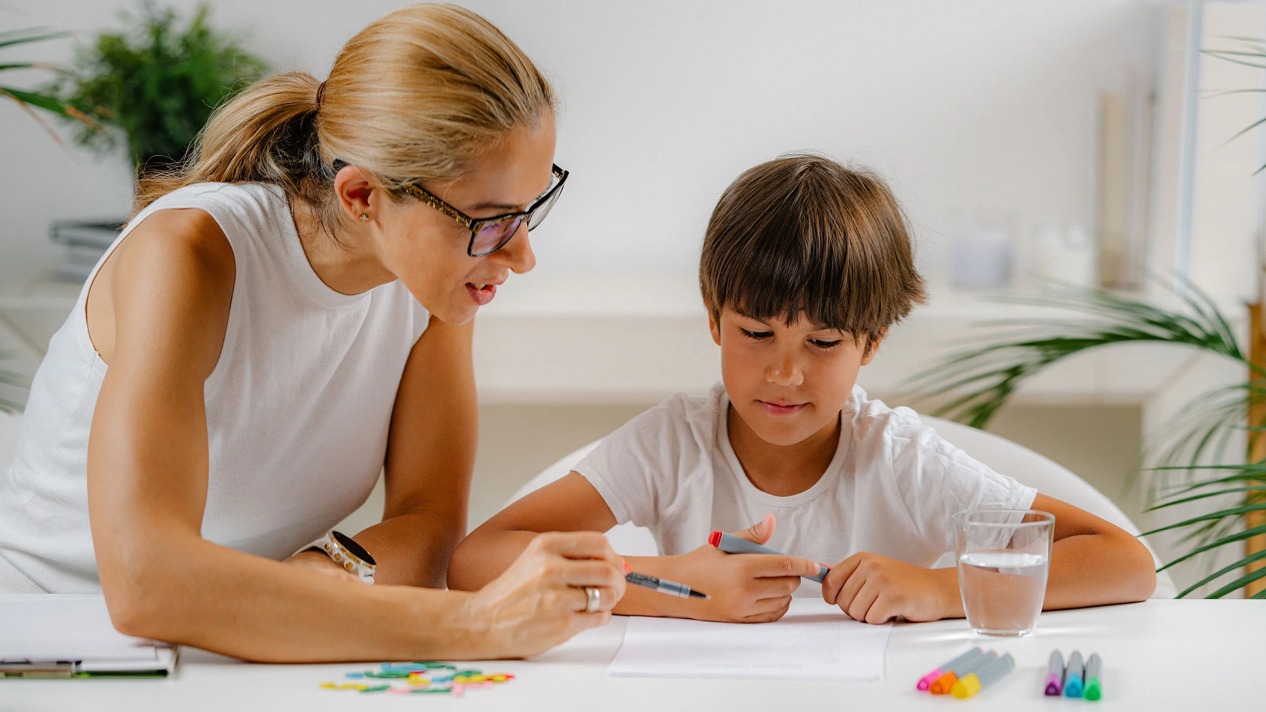 A female therapist wearing glasses guides a young boy during a drawing activity at a white table.