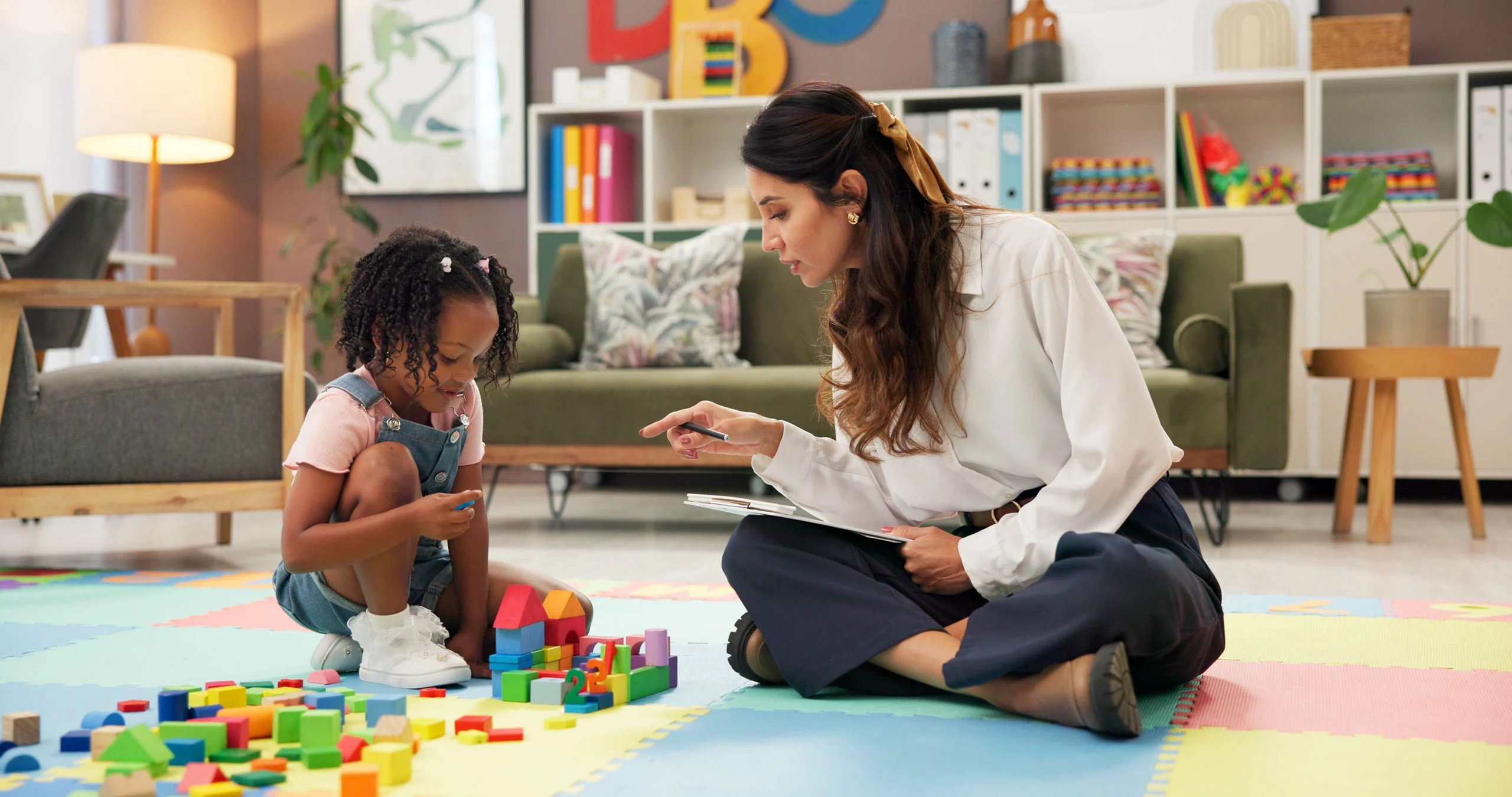 A female specialist with a clipboard sitting cross-legged on a colorful floor mat, interacting with a young girl building with wooden blocks.