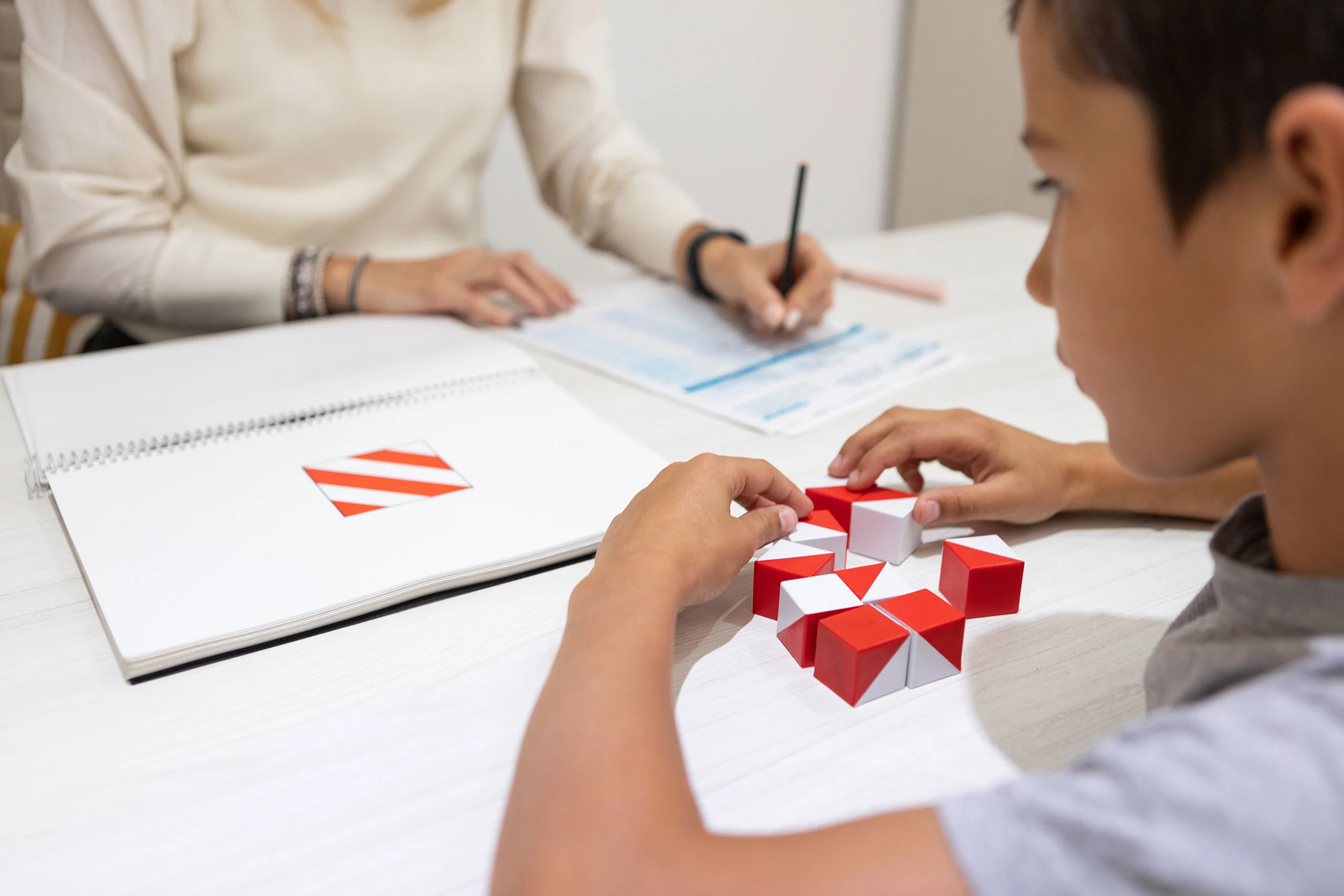 A child completes a Kohs block design test by arranging red and white cubes while an evaluator fills out a scoring form.