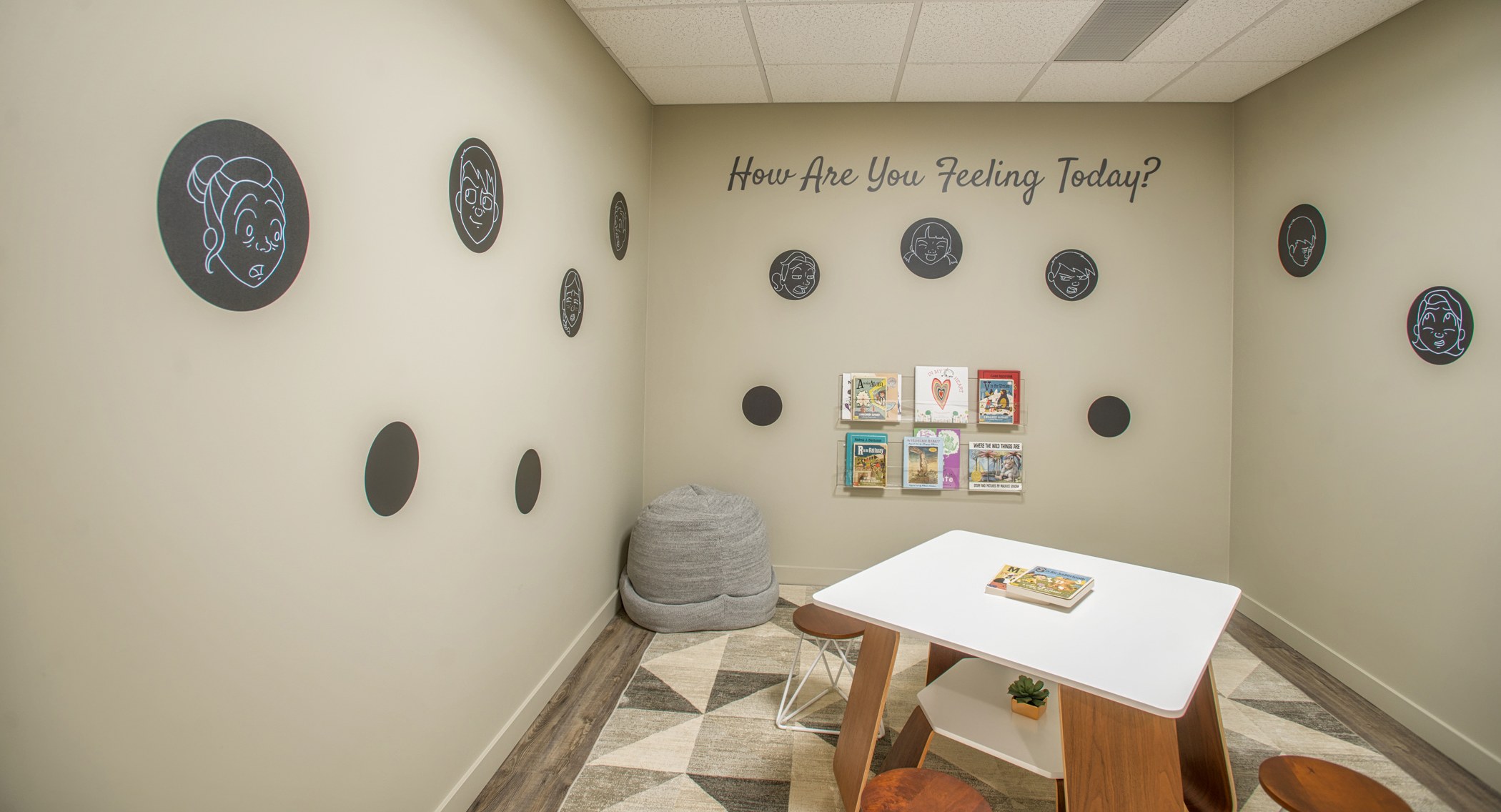 A children's therapy room with a wall decal reading 'How Are You Feeling Today?' surrounded by circular emoji illustrations, a small activity table, a beanbag chair, and a book display.